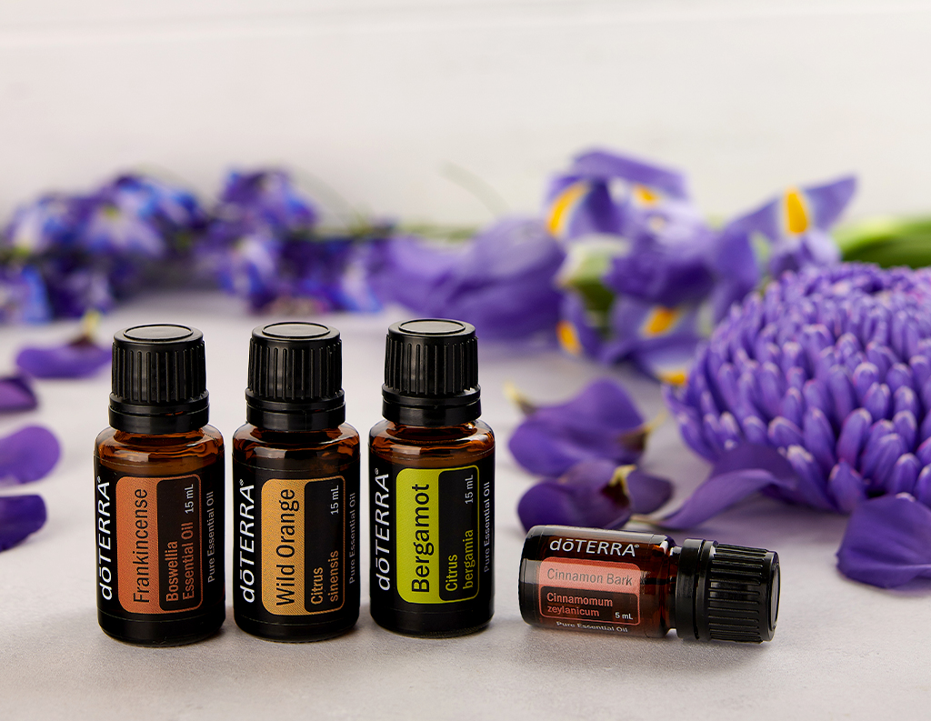 The image features four doTERRA essential oil bottles placed in a row, with a backdrop of purple flowers. The oils include Frankincense (Boswellia Essential Oil, 15 mL), Wild Orange (Citrus sinensis, 15 mL), Bergamot (Citrus bergamia, 15 mL), and Cinnamon Bark (Cinnamomum zeylanicum, 5 mL). The vibrant purple flowers and petals in the background add a serene and natural touch, complementing the oils' earthy and citrusy tones.