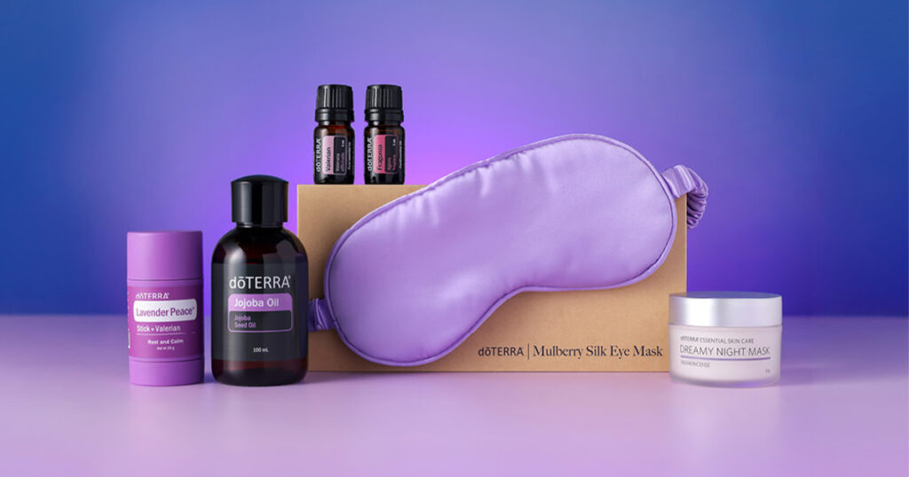 Collection of doTERRA wellness products arranged on a purple background, including a Lavender Peace stick, Jojoba Oil bottle, two essential oil bottles, a Mulberry Silk Eye Mask in a box, and a jar of Dreamy Night Mask.