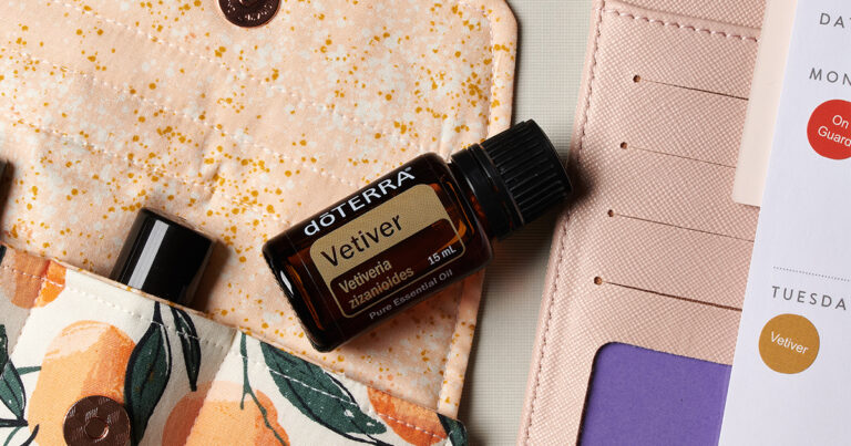 Close-up of a doTERRA Vetiver essential oil bottle placed on a patterned fabric pouch with peach and orange accents, next to a planner page with a schedule indicating 'Vetiver' usage on Tuesday.