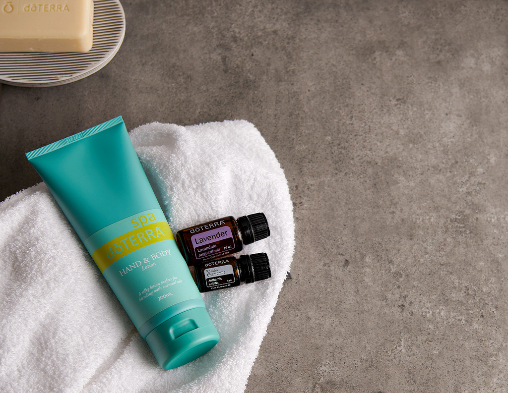 This image shows a collection of spa products on a white towel, laid out on a gray stone surface. The products include a tube of dōTERRA Spa Hand & Body Lotion, along with two essential oil bottles—one labeled Lavender and the other Roman Chamomile. In the background, there is a bar of dōTERRA soap placed on a striped soap dish. The overall setup suggests a relaxing, self-care atmosphere.