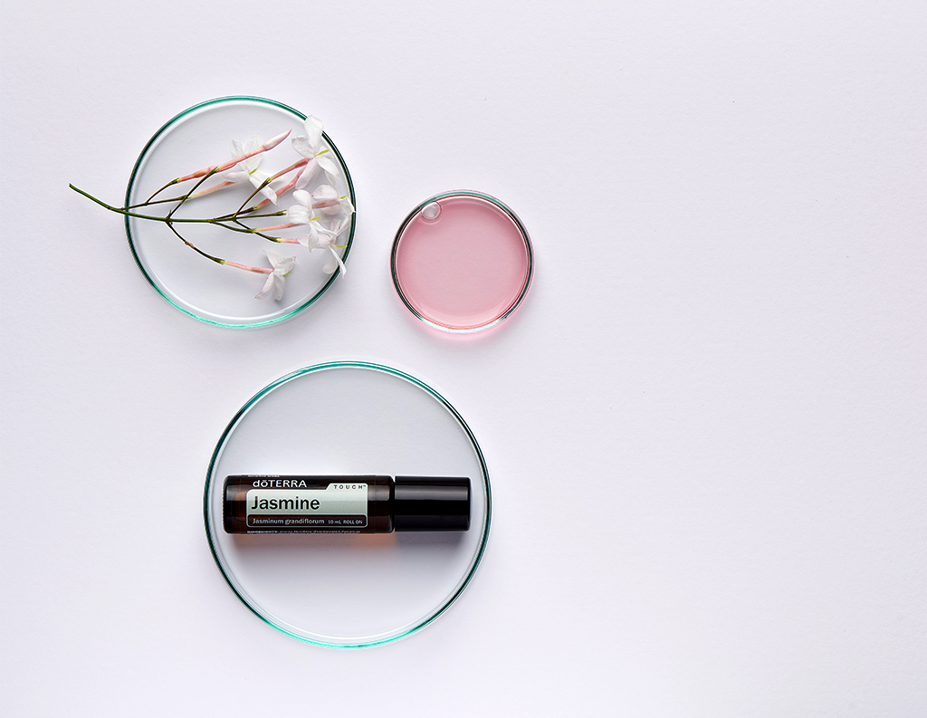 The image shows a doTERRA Jasmine Touch roller bottle, labeled Jasminum grandiflorum, placed in a glass dish. Surrounding it are two other glass dishes, one containing a sprig of delicate jasmine flowers and the other filled with a light pink liquid. The scene is set against a clean, minimalist white background, highlighting the purity and elegance of jasmine.