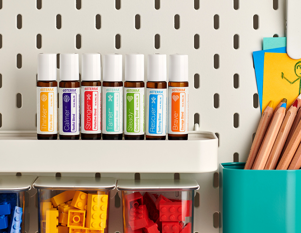 The image shows seven roller bottles from doTERRA's Kids Collection arranged on a shelf. Each bottle has a colorful label, representing different blends: Thinker (Focus Blend), Calmer (Restful Blend), Stronger (Protective Blend), Tamer (Digestive Blend), Steady (Grounding Blend), Rescuer (Soothing Blend), and Brave (Courage Blend). Below the shelf, there are clear bins filled with colorful LEGO bricks, and on the side, a teal container holds pencils and papers, suggesting a playful and creative space designed for children.