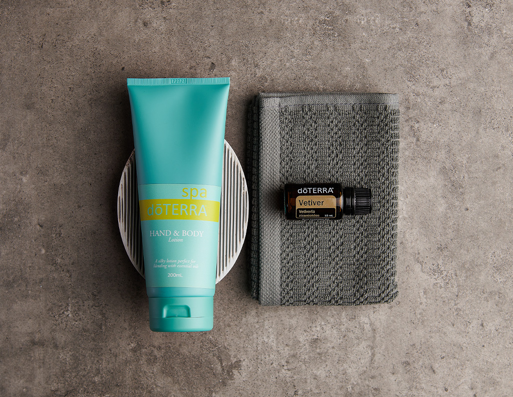 The image displays a tube of doTERRA Spa Hand & Body Lotion (200 mL) alongside a bottle of doTERRA Vetiver essential oil (Vetiveria zizanioides, 15 mL). The lotion rests on a circular dish, while the Vetiver oil is placed on a folded gray towel, all set against a textured gray surface. The arrangement evokes a calm, spa-like atmosphere, suggesting a combination of skincare and aromatherapy.