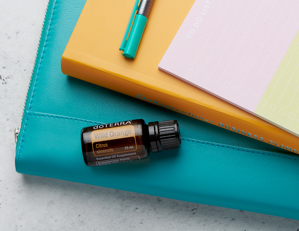 The image shows a 15 mL bottle of doTERRA Wild Orange essential oil labeled Citrus sinensis placed on top of a turquoise folder. Next to it, there is a yellow business planner, a to-do list pad, and a turquoise pen. The scene suggests a workspace setting, with the Wild Orange oil possibly being highlighted for its energizing or refreshing properties.