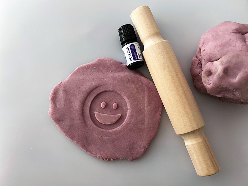 This image shows a crafting scene featuring lavender-colored playdough, a wooden rolling pin, and a bottle of doTERRA Calmer essential oil. The playdough has been rolled flat with an imprint of a smiling face stamped into it.