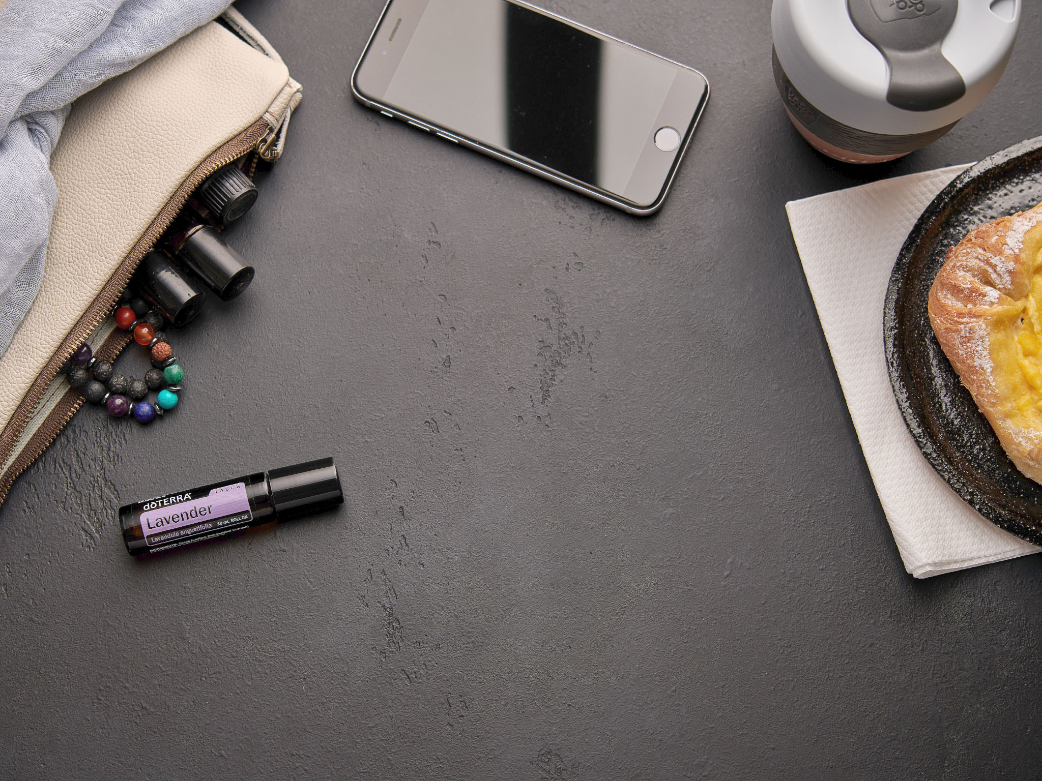 The image displays a flat lay of a desktop scene featuring a roll-on lavender essential oil bottle, a beige pouch with small vials peeking out, a beaded bracelet, a smartphone, a reusable cup, and a pastry on a black plate with a napkin. The setting suggests relaxation or a moment of self-care, combining natural remedies with a snack and drink.