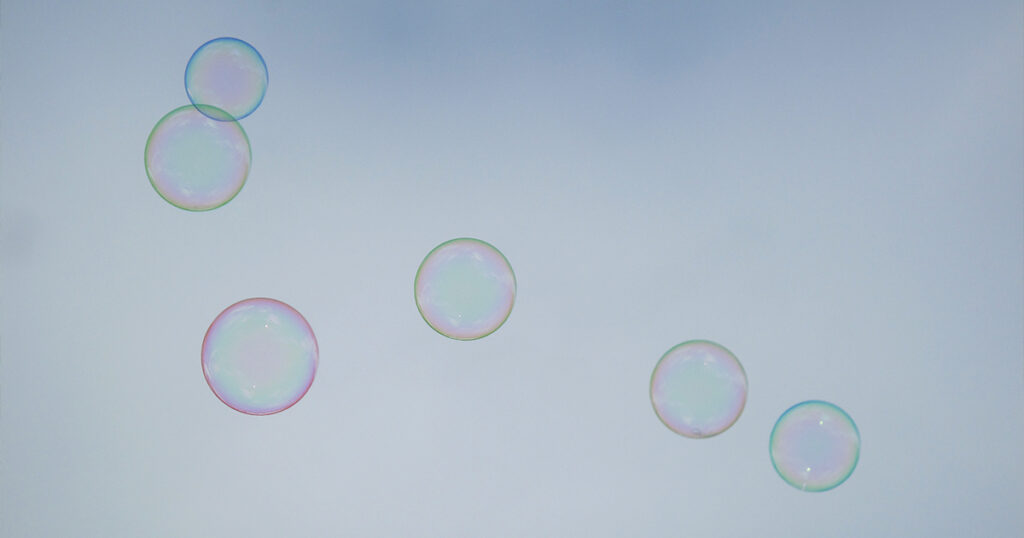 The image shows a group of six delicate soap bubbles floating against a soft, overcast sky. The bubbles have a translucent quality with subtle, iridescent hues of pink, green, blue, and purple reflecting on their surfaces, creating a gentle, whimsical effect.