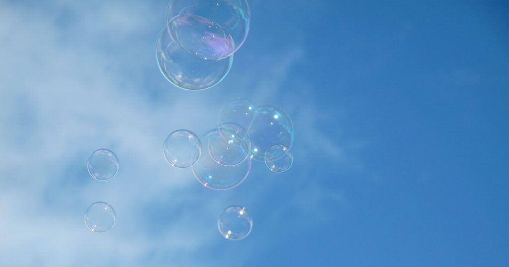 The image shows a cluster of translucent soap bubbles drifting upward against a bright blue sky, with a few wispy white clouds in the background. The bubbles have a subtle rainbow sheen, reflecting soft hues of pink, blue, and green, creating a playful, airy atmosphere.