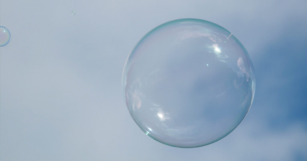 A close-up of a single, iridescent soap bubble floating against a soft, blue-gray sky. The bubble reflects hints of pink and white from the surrounding light, creating a delicate, otherworldly effect. Faint traces of smaller bubbles are visible in the background, blending seamlessly into the cloudy atmosphere.