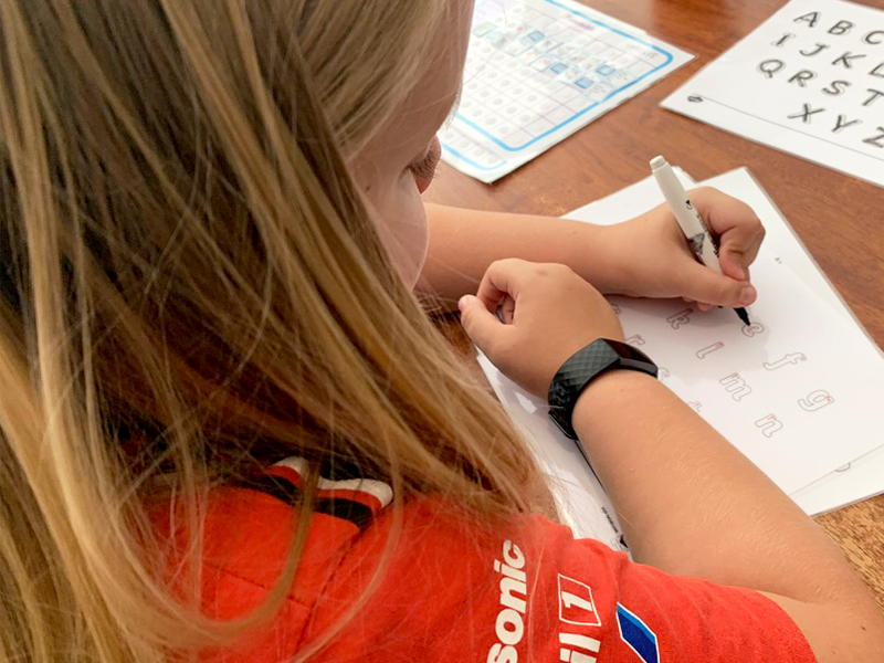 A young boy with long blonde hair, wearing a red Panasonic racing shirt, practices handwriting at a wooden table. He traces lowercase letters on a worksheet with a black marker, while an uppercase alphabet guide and a laminated alphabet chart are visible nearby on the table.