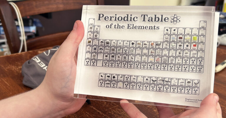 The image shows a clear acrylic periodic table display being held by two hands. The table is labeled "Periodic Table of the Elements" with a small atomic symbol graphic beside it. Each element block includes its atomic number, symbol, and a small piece of the actual element or a representative image for more dangerous or rare elements. The acrylic sheet appears to be high-quality, produced by "Engineered Labs Protoshop LLC," as indicated in the bottom right corner. The table is resting against a wooden surface, with a gray drawstring bag partially visible in the background.