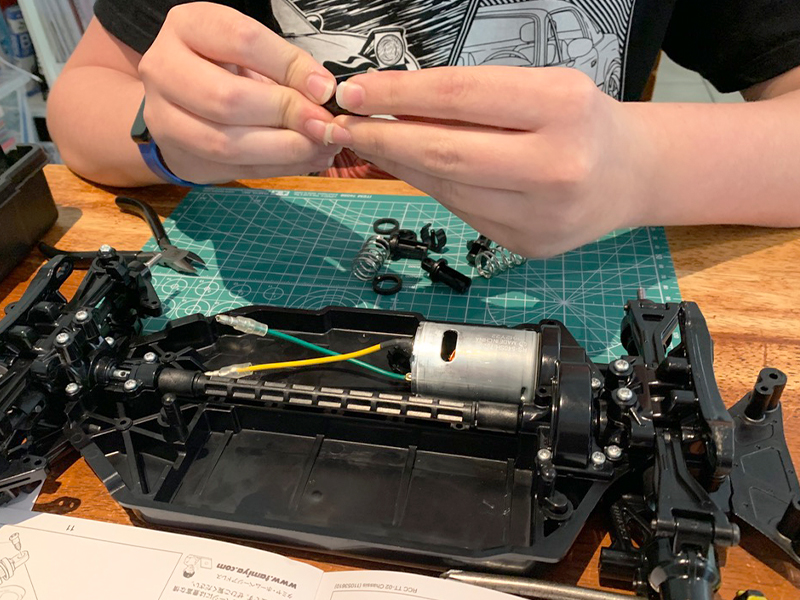 A person assembles a remote-controlled car chassis on a wooden table, carefully holding small components with both hands. The partially assembled RC car reveals its inner wiring and motor, surrounded by tools, springs, and black plastic parts atop a green cutting mat.