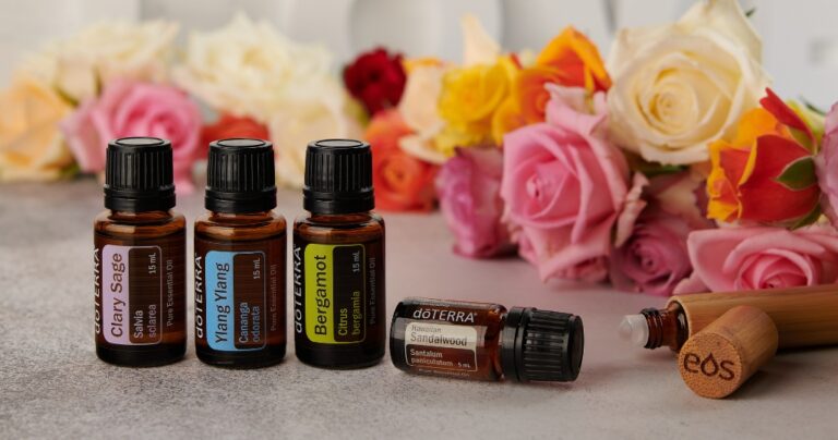 Four doTERRA essential oil bottles—Clary Sage, Ylang Ylang, Bergamot (all 15 mL), and Hawaiian Sandalwood (5 mL)—are arranged on a stone surface, with the Sandalwood bottle lying on its side next to a bamboo roller labeled “eos.” In the background, a lush arrangement of roses in pink, yellow, and red adds a vibrant floral touch.