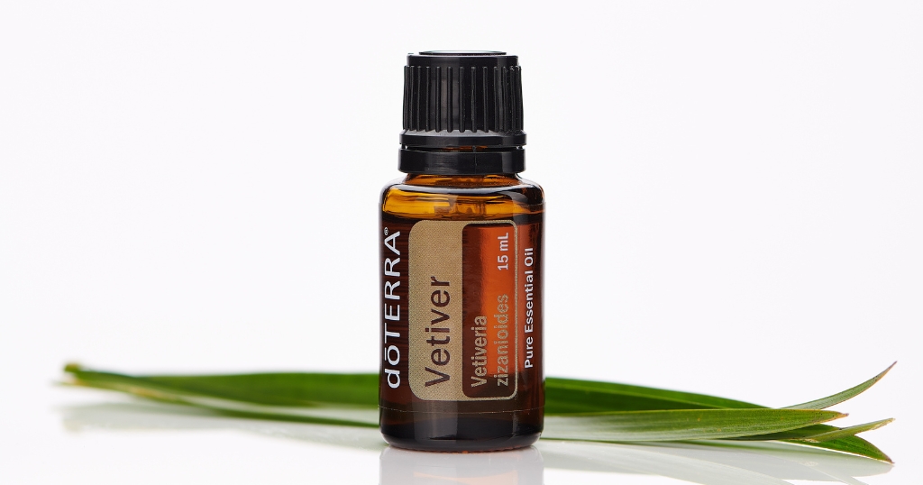 A 15 mL amber bottle of doTERRA Vetiver essential oil stands upright on a white surface, surrounded by slender green vetiver leaves. The label reads "Vetiveria zizanioides" and "Pure Essential Oil," highlighting its earthy botanical source.