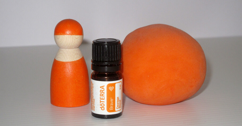 An orange ball of aromatherapy playdough sits beside a small bottle of doTERRA Brave essential oil blend and an orange Grapat Nin - a wooden play figure with a spherical head and brightly colored body, used for sensory and imaginative play."