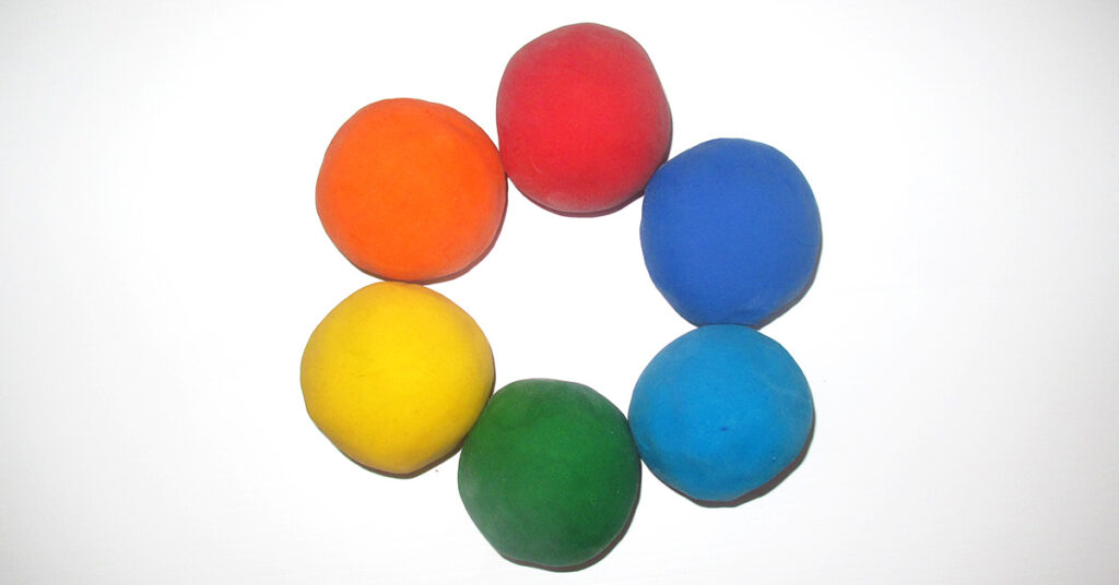 Six balls of aromatherapy playdough arranged in a circle on a white background, in red, orange, yellow, green, light blue, and dark blue, showing the rainbow colors often used for sensory play with aromatherapy playdough.
