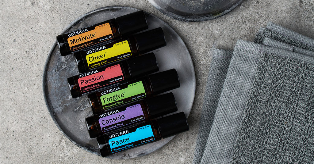 Flat lay of six doTERRA Touch essential oil rollers arranged on a gray stone plate. Each bottle is labeled with a mood-related name: Motivate (orange), Cheer (yellow), Passion (red), Forgive (green), Console (purple), and Peace (blue). A second plate 
 sits above them, next to neatly folded gray towels, all on a concrete surface.
