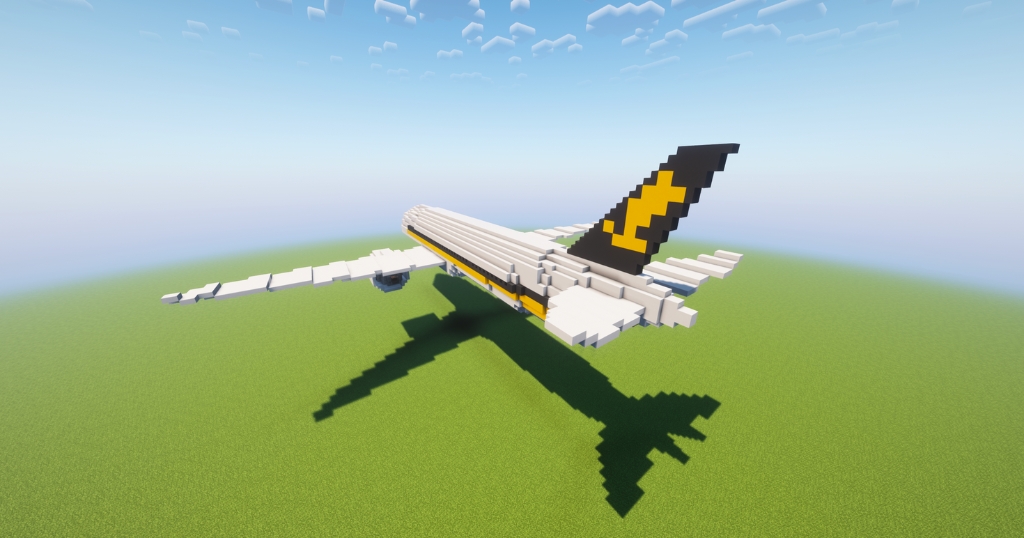 A Minecraft-style Boeing 777-200ER airplane built with white, yellow, and black blocks flies over a flat grassy landscape under a bright blue sky. The airplane features a distinctive black tail fin with a bold yellow arrow-like design, casting a detailed shadow on the terrain below.