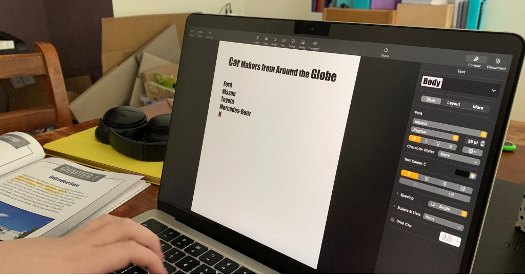 A student works on a laptop, creating a document titled "Car Makers from Around the Globe" with a list including Ford, Nissan, Toyota, and Mercedes-Benz. A textbook open to a chapter introduction and a pair of black headphones rest on the desk nearby, suggesting a home study setup.