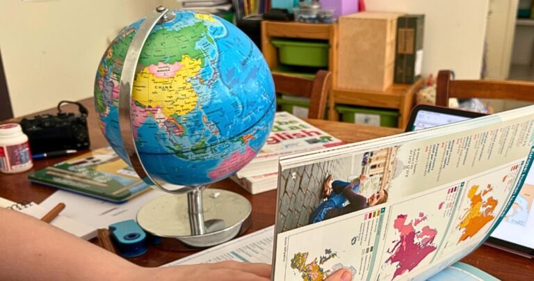 Colorful educational setup featuring a world globe, geography workbook, and learning materials spread across a table. A student holds a book open to a page with maps and a travel photo, reinforcing a hands-on approach to create your own unit studies with engaging, real-world content.