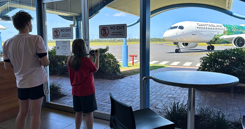 Two children stand inside an airport terminal, watching a QantasLink airplane through large glass doors marked “NO EXIT.” One child is filming the plane with a small camera while the other looks on, and the aircraft outside features a vivid green and white livery.
