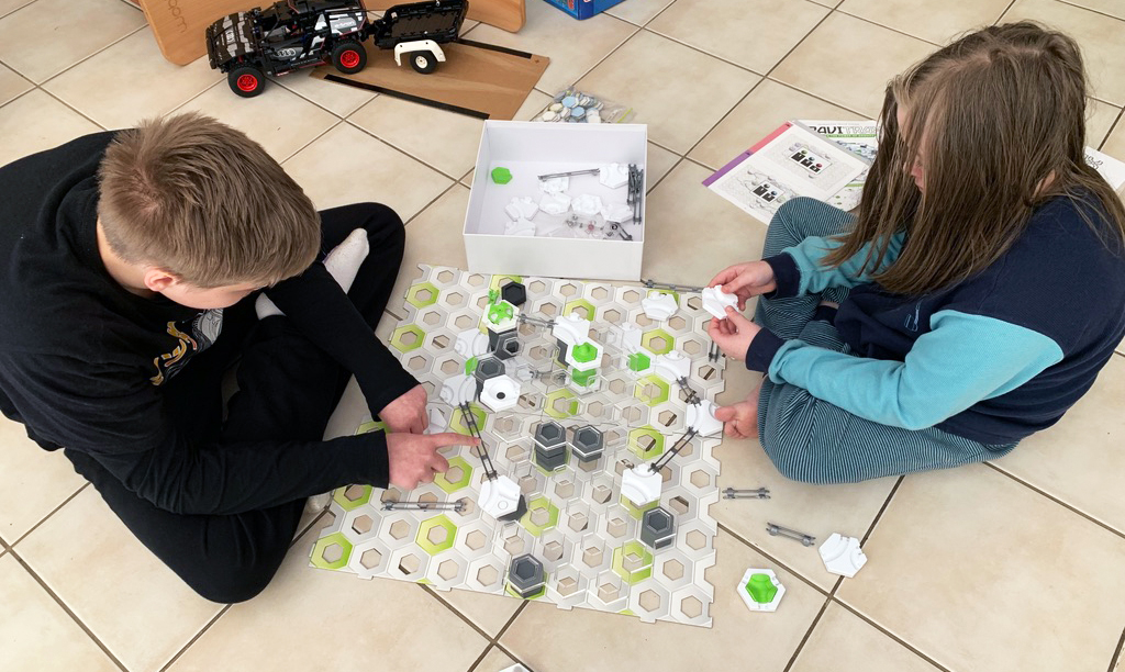 Two children sit on a tiled floor building a marble run game with hexagonal pieces, ramps, and towers spread out between them. Instruction sheets and boxes are nearby as they work together on the STEM-style project. The image reflects hands-on learning and collaboration in a neurodivergent homeschooling environment.