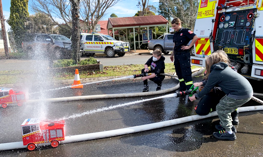 Children participate in a hands-on activity, spraying water from fire hoses alongside firefighters near a fire truck. Small red toy fire engines and safety cones line the wet pavement under clear skies. The image highlights experiential learning and community engagement in neurodivergent homeschooling.