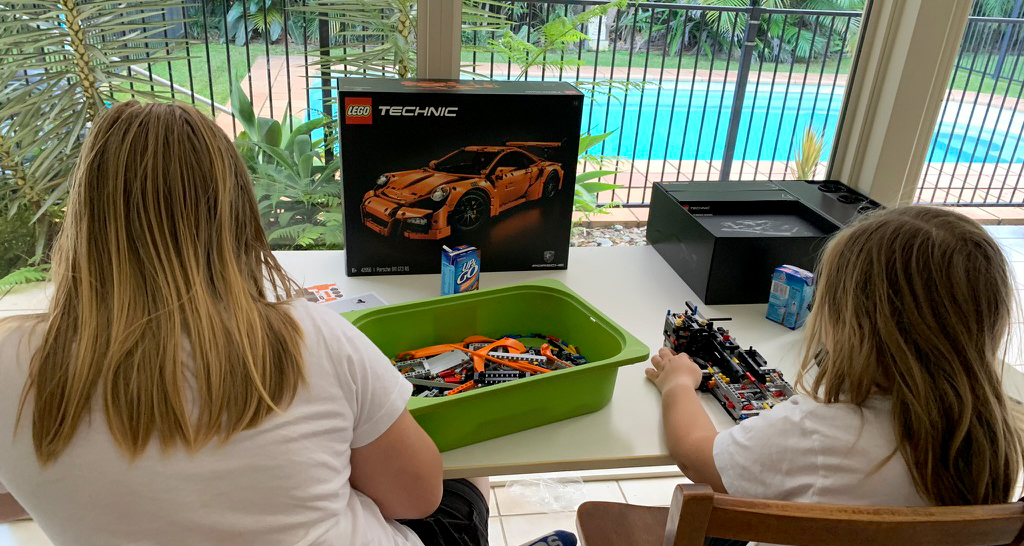 Two children sit at a table building a LEGO Technic car set together, with a green bin of pieces in front of them and the LEGO box showing an orange sports car. They are working indoors beside large windows overlooking a swimming pool and lush garden. The scene reflects a hands-on homeschooling activity focused on creativity and engineering.