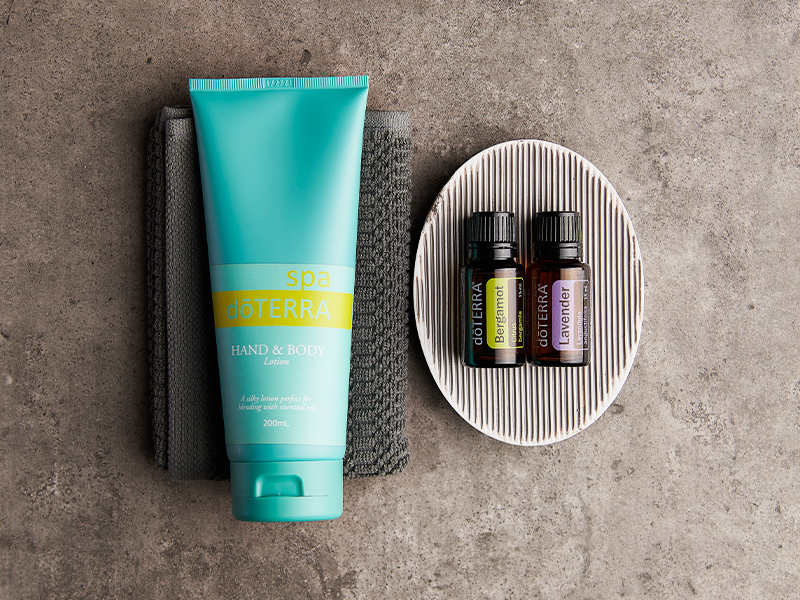 Flat lay of a teal doTERRA Hand & Body Lotion tube placed on folded towels beside a small ribbed dish holding two essential oil bottles labeled Bergamot and Lavender. The minimalist spa-like setup introduces basic elements in essential oils 101, emphasizing simple, soothing self-care.