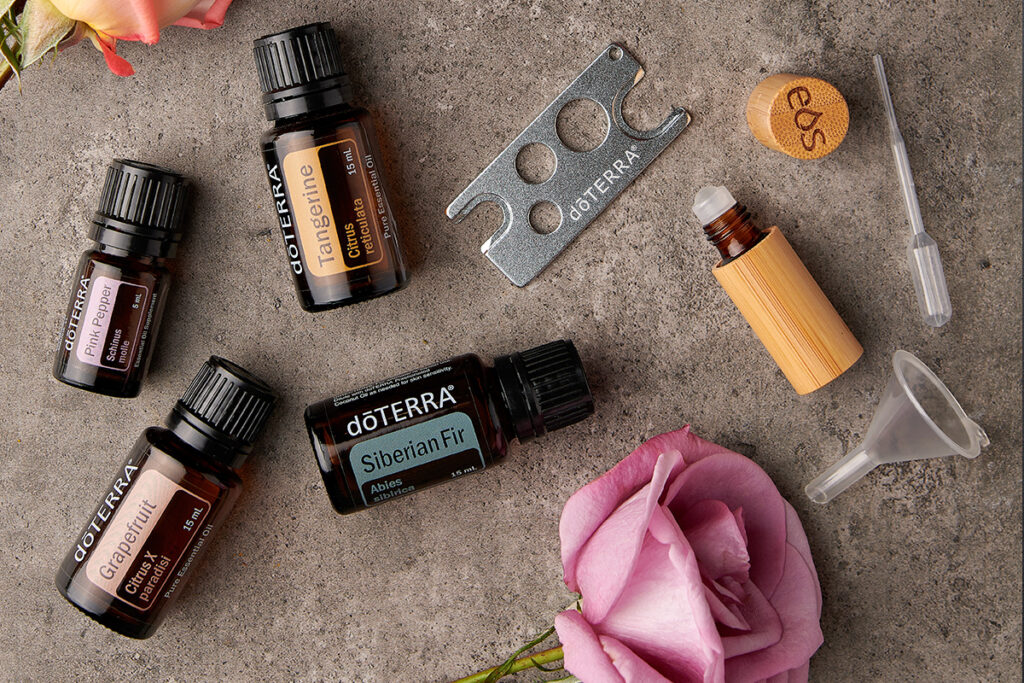A flat lay of doTERRA essential oils including Pink Pepper, Grapefruit, Tangerine, and Siberian Fir, surrounded by blending tools like a pipette, mini funnel, oil key, and a bamboo roller bottle. Scattered rose petals and flowers add a soft touch, illustrating a setup for creating DIY blends at home.