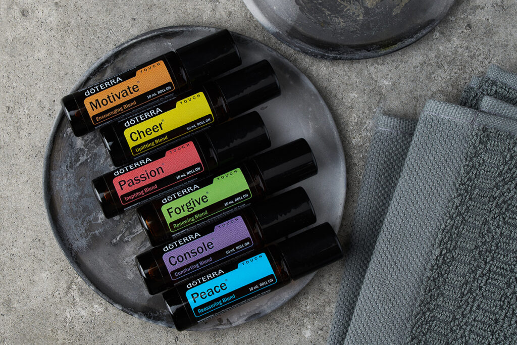 Flat lay of six doTERRA Touch essential oil rollers arranged on a gray stone plate. Each bottle is labeled with a mood-related name: Motivate (orange), Cheer (yellow), Passion (red), Forgive (green), Console (purple), and Peace (blue). A second plate
sits above them, next to neatly folded gray towels, all on a concrete surface.