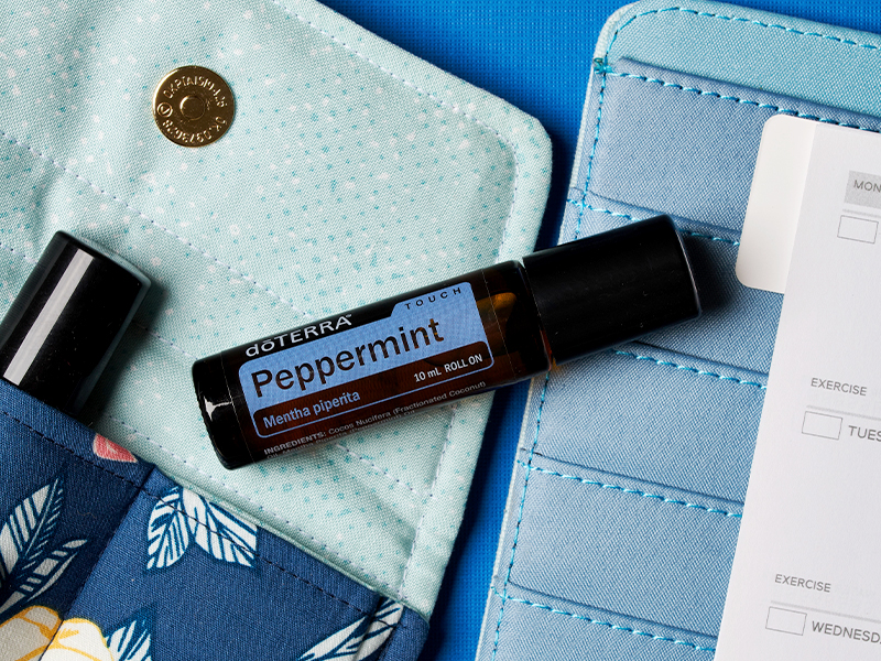 A doTERRA Touch Peppermint roller bottle rests on a soft teal pouch beside a wallet and planner, emphasizing its convenience for on-the-go use. As a prediluted hot oil, this peppermint blend removes the guesswork, offering cooling relief without the need to measure carrier oils.