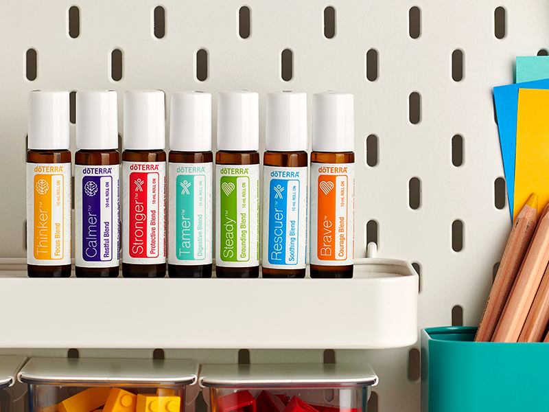 Seven doTERRA Kid’s Collection essential oil rollers - Thinker, Calmer, Stronger, Tamer, Steady, Rescuer, and Brave - are neatly lined up on a white shelf against a pegboard wall, next to colorful paper and pencils. These kid-safe oils are designed with gentle blends for focus, relaxation, immunity, digestion, and emotional support.