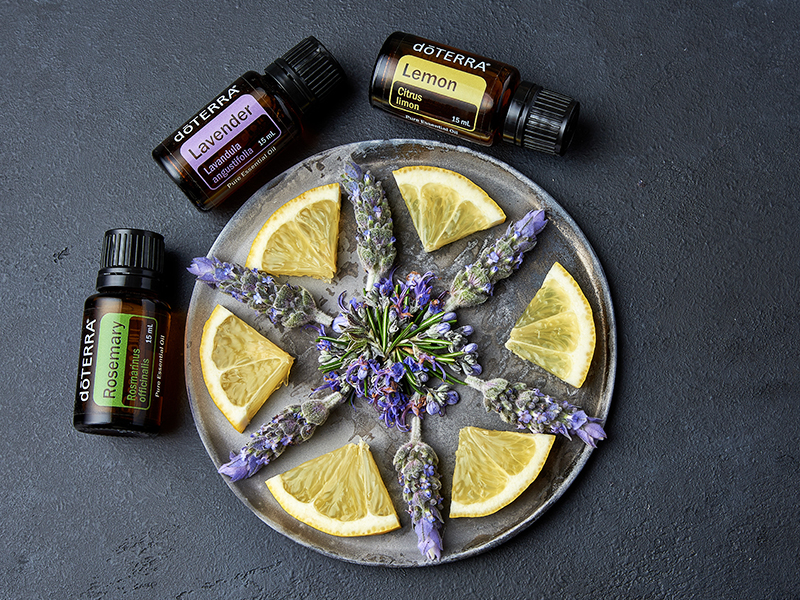 Flat lay of a metal plate arranged with alternating slices of lemon and sprigs of lavender, centered around a small lavender bouquet, next to three doTERRA essential oil bottles labeled Lavender, Lemon, and Rosemary. The dark slate background contrasts with the bright citrus and florals, highlighting the natural ingredients used in essential oils.