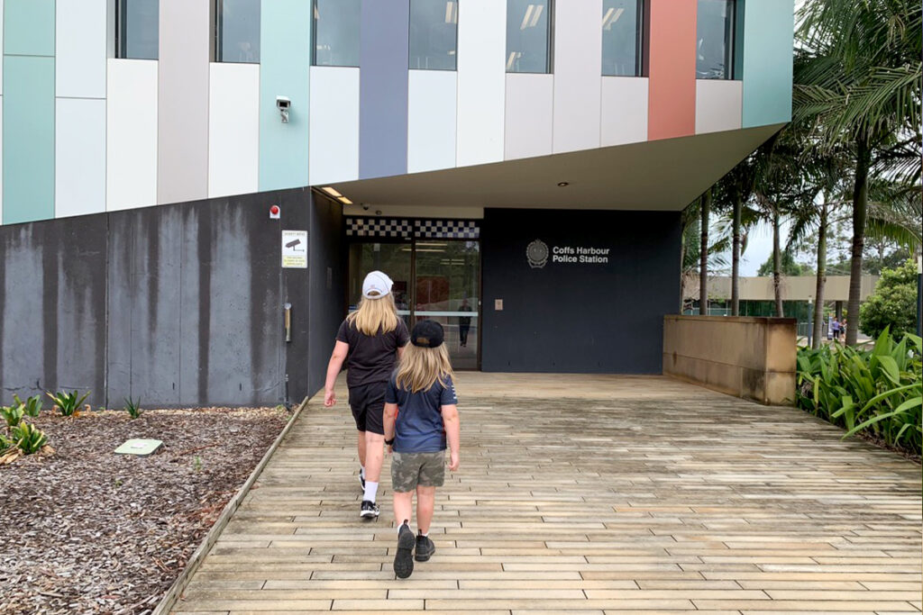 Two children walking toward the entrance of the Coffs Harbour Police Station, a modern building with colorful vertical panels and a wood-plank pathway. This hands-on homeschool project offers a real-world learning experience about law enforcement and community roles.