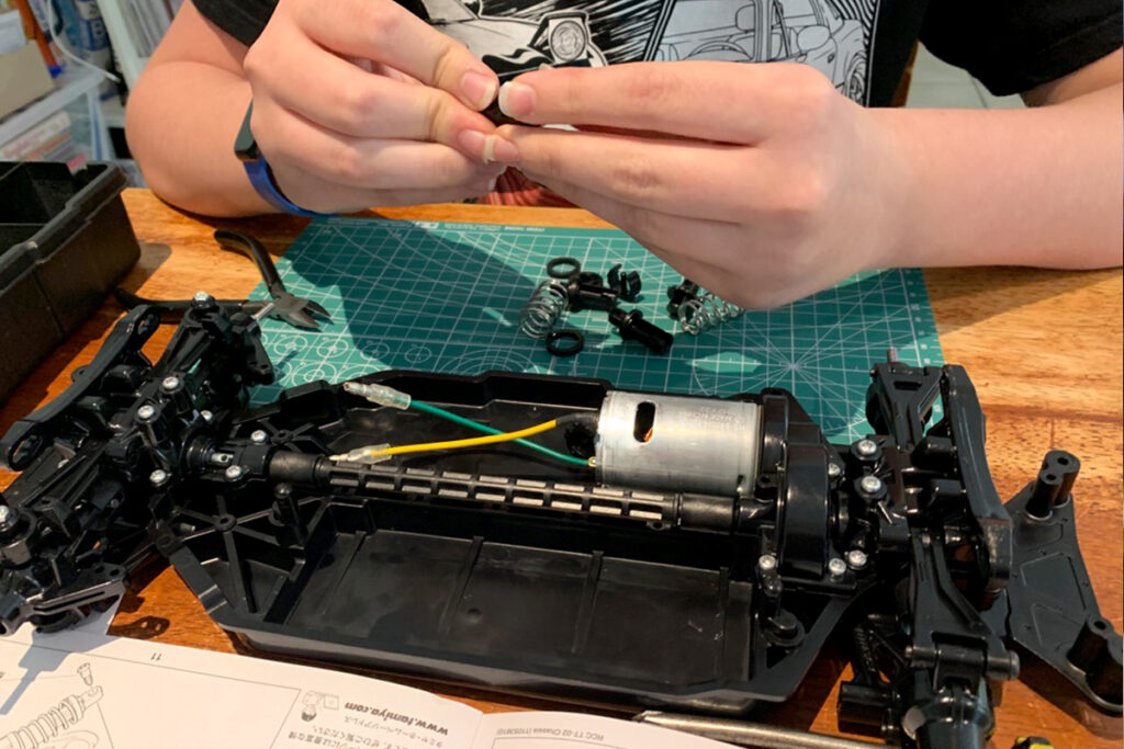 Child assembling parts of a remote-controlled car on a wooden table with a green cutting mat, surrounded by small components like springs and screws. This hands-on homeschool project encourages STEM learning through mechanical construction.