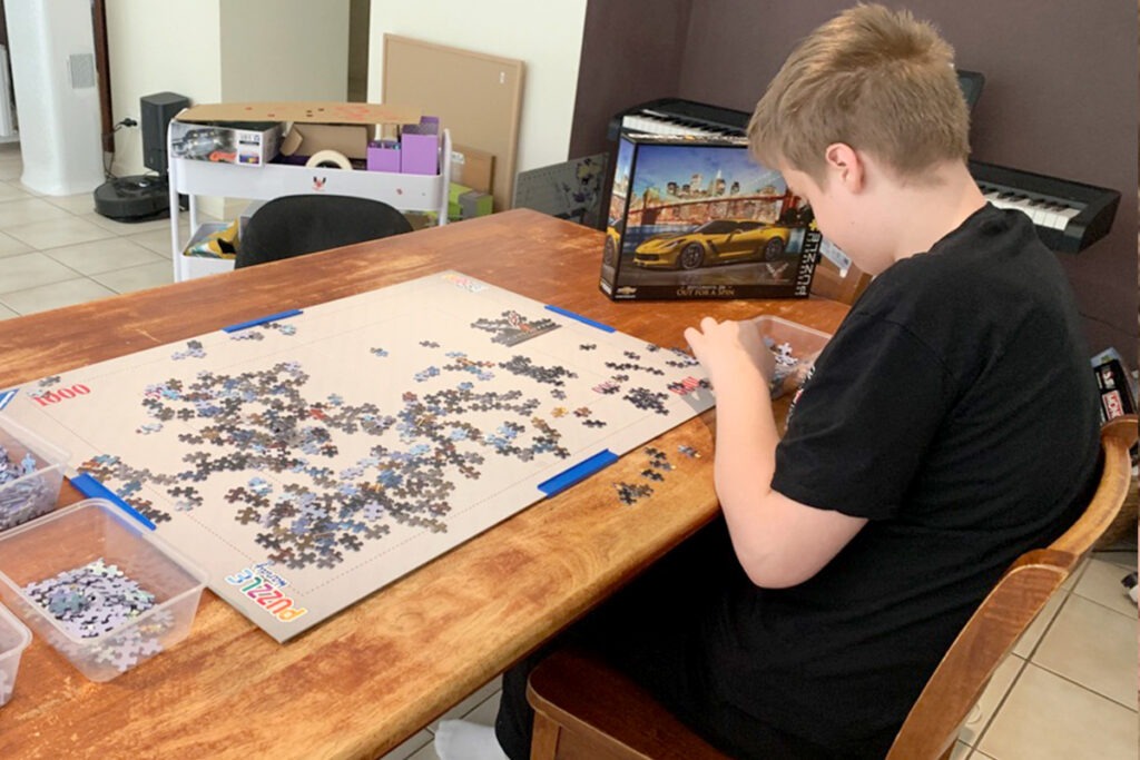 Child focused on assembling a large jigsaw puzzle at the dining table, with sorted pieces in plastic bins and the puzzle box showing a yellow Corvette. A quiet, screen-free moment that fits naturally into a relaxed homeschool rhythm supporting focus and executive function skills.