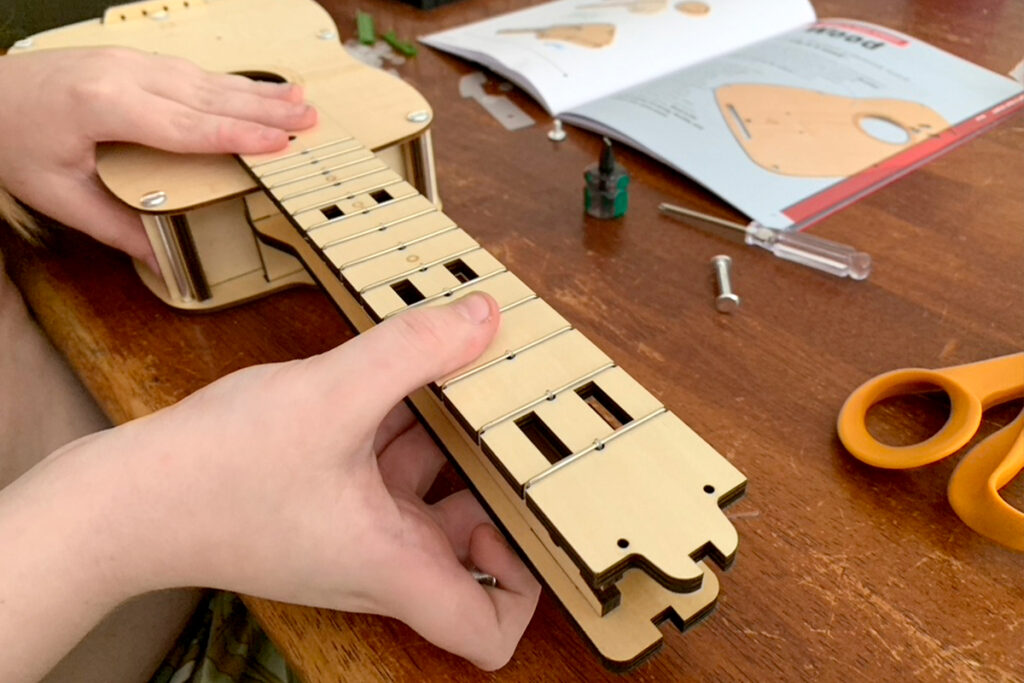 Child building a wooden ukulele from a Kiwi Crate kit, hands assembling the fretboard while following illustrated instructions on a cluttered table. A calming, hands-on activity as part of a flexible homeschool schedule for a child with ADHD and PDA.