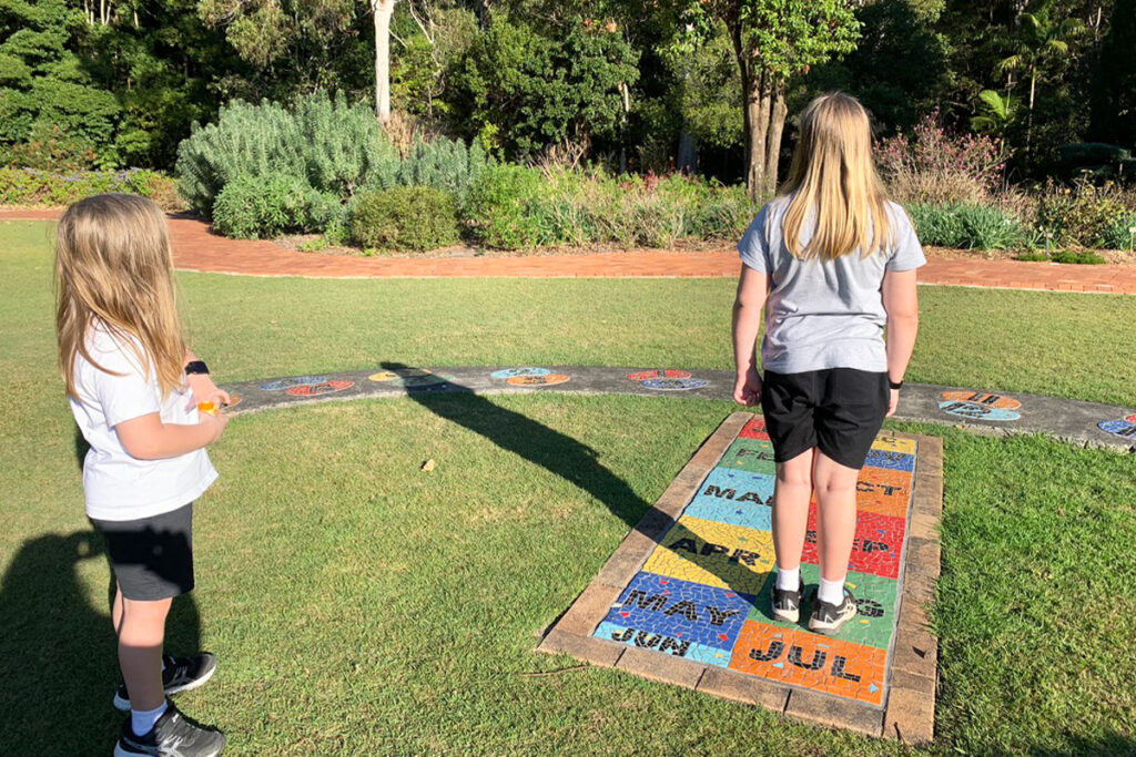 Two boys stand on a grassy outdoor path decorated with colorful mosaic tiles labeled with months of the year. This sensory-rich environment supports ADHD homeschooling by combining movement, visual cues, and nature-based learning.