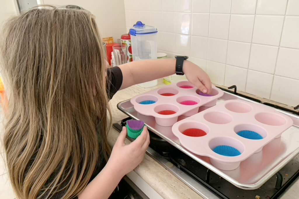 Child with long hair sets paper cupcake cases into pink silicone muffin trays on a baking sheet - a real-life example of what counts as learning.