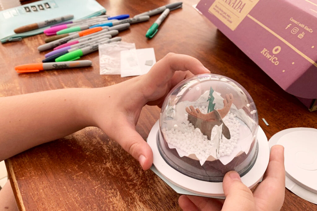 Hands assembling a homeschool project - a mini snow globe-style dome with paper cut-outs and foam beads, surrounded by art supplies.