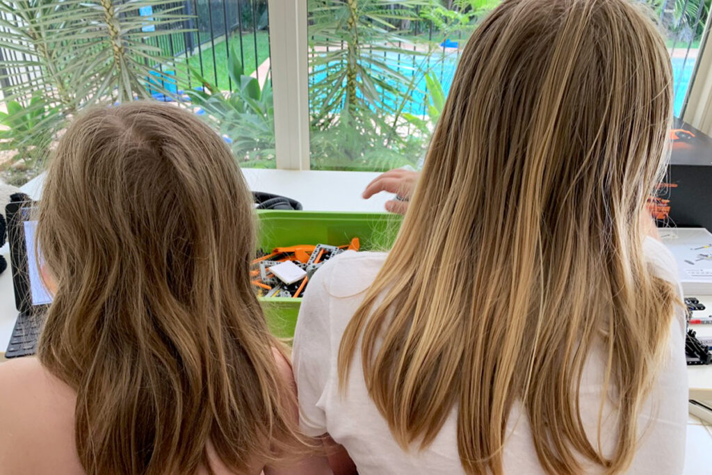 Two boys with long hair sit side by side at a table indoors, focused on a tray of LEGO robotics parts with a laptop nearby. As part of their neurodivergent homeschooling, they're engaging in hands-on STEM learning in a calm, sensory-friendly environment overlooking a pool and garden.