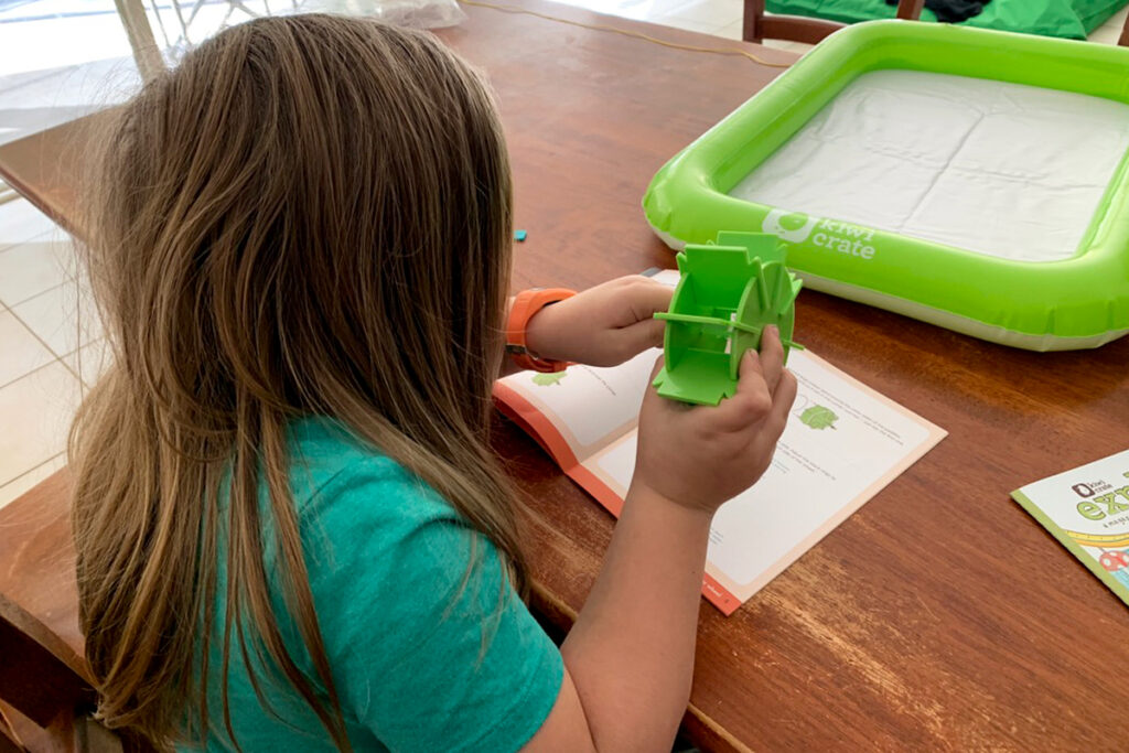 Child with long hair wearing a teal shirt sits at a wooden table assembling a green plastic water wheel from a KiwiCo kit, following illustrated instructions. Hands-on science kits like this are often used in homeschooling autism environments to support sensory learning and STEM exploration.