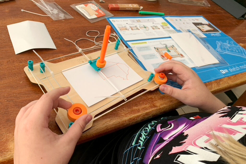 A child’s hands adjust a DIY drawing machine that’s tracing a map on paper in a neurodivergent homeschooling activity.