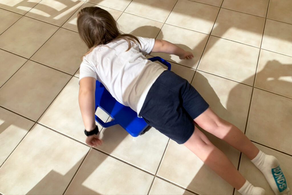 A young child lies on their stomach on a blue scooter board, using their arms to move across a tiled floor. The sunlight casts shadows from nearby furniture, creating a playful environment that reflects sensory-friendly homeschooling activities for neurodivergent kids.
