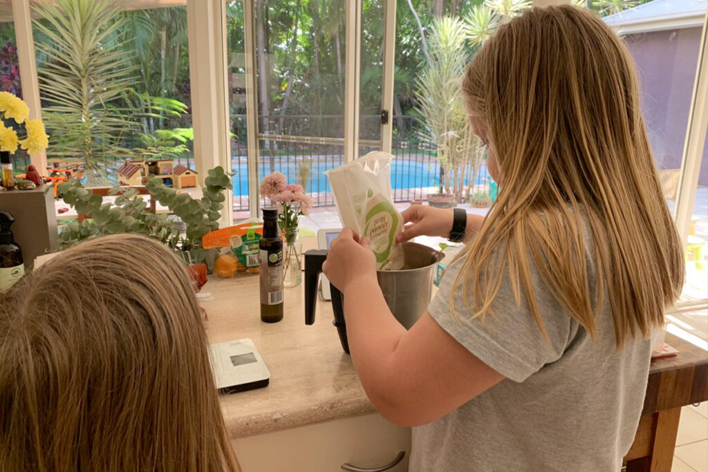 Two children in a sunlit kitchen preparing food as part of an interest-led homeschooling activity. One child is pouring organic flour into a mixing bowl while the other watches, with fresh ingredients, a kitchen scale, and flowers on the counter beside them. A backyard pool and lush greenery are visible through large windows.
