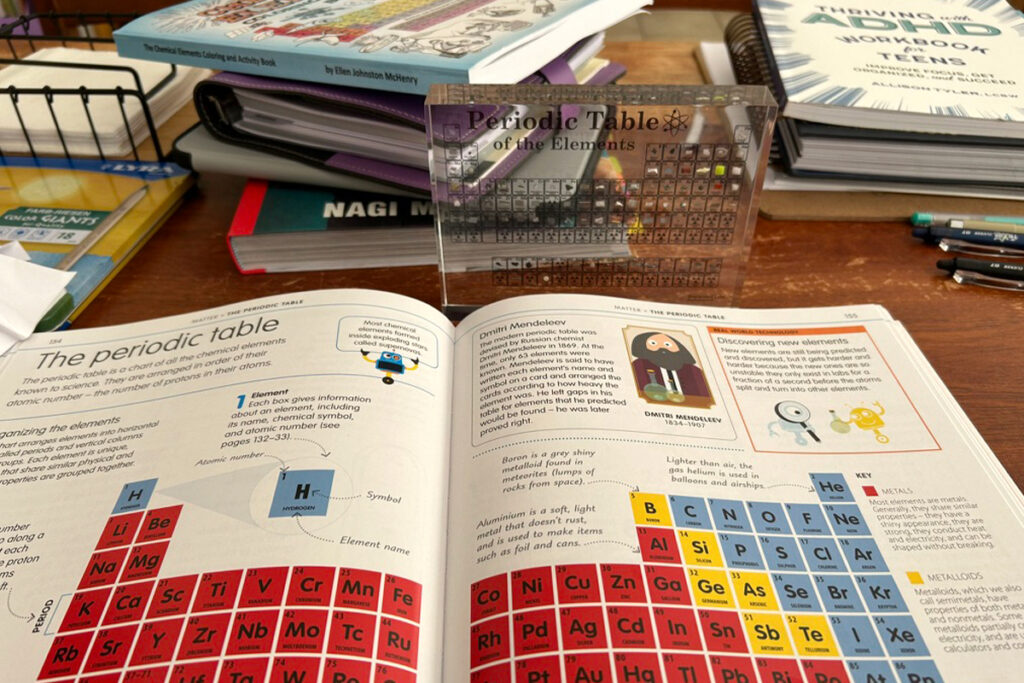 Open science workbook spread to a lesson titled The periodic table with colorful element blocks and a section on Dmitri Mendeleev. A clear acrylic Periodic Table of the Elements chart stands behind the book among stacked workbooks and pencils on a desk. A homeschooling curriculum science setup focused on learning the periodic table.