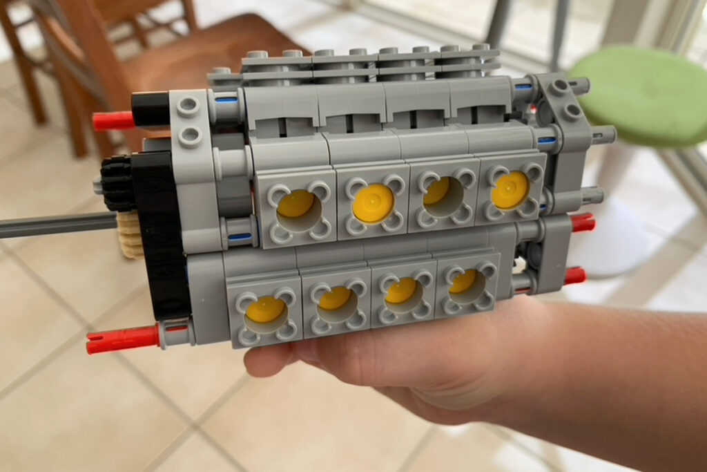 A hand holds a detailed LEGO Technic engine build made from gray and yellow pieces with red axle pins sticking out. The close up view highlights the layered structure and moving parts, illustrating self directed learning through hands on mechanical building and experimentation.