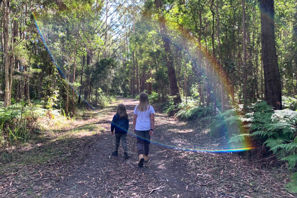 wo children walk along a quiet forest trail surrounded by tall trees and greenery with sunlight creating a rainbow lens flare across the path. The scene reflects a calm outdoor learning moment in neurodivergent homeschooling, offering an alternative to traditional settings where a child might feel like they are falling behind.