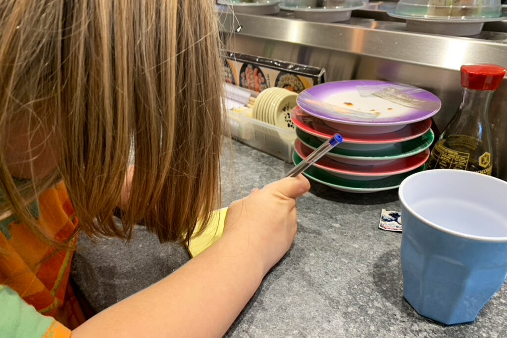 Child at a sushi train counter holds a pen and points toward a stack of color coded plates while counting and comparing them. Photo illustrates homeschooling a child with pathological demand avoidance by turning sushi train pricing into pressure free maths and bill estimating.